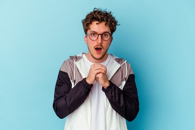 Young caucasian man wearing eyeglasses isolated on blue background praying for luck, amazed and opening mouth looking to front.