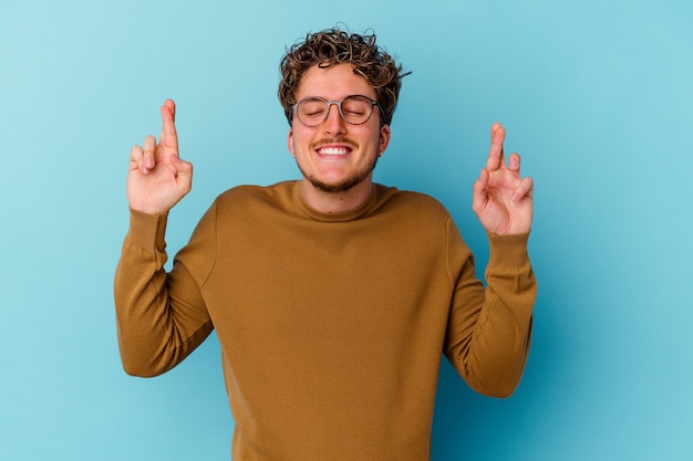 Young caucasian man wearing eyeglasses isolated on blue wall crossing fingers for having luck