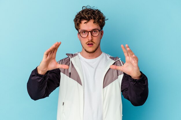 Young caucasian man wearing eyeglasses isolated on blue wall holding something with palms, offering to the front