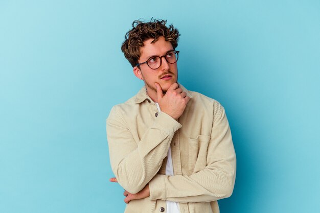 Young caucasian man wearing eyeglasses isolated on blue wall thinking and looking up, being reflective, contemplating, having a fantasy.