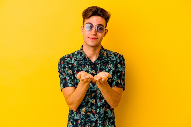 Young caucasian man wearing a Hawaiian shirt isolated on yellow wall holding something with palms