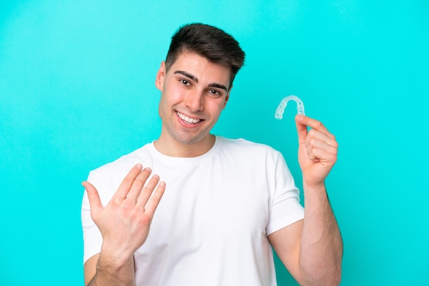 Young caucasian man wearing holding invisible braces isolated on blue background inviting to come with hand Happy that you came