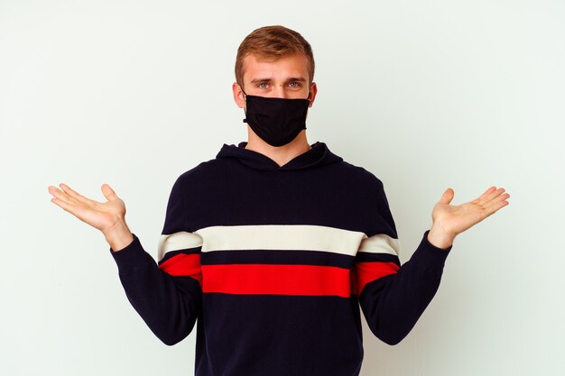 Young caucasian man wearing a mask for virus isolated on white background makes scale with arms, feels happy and confident.