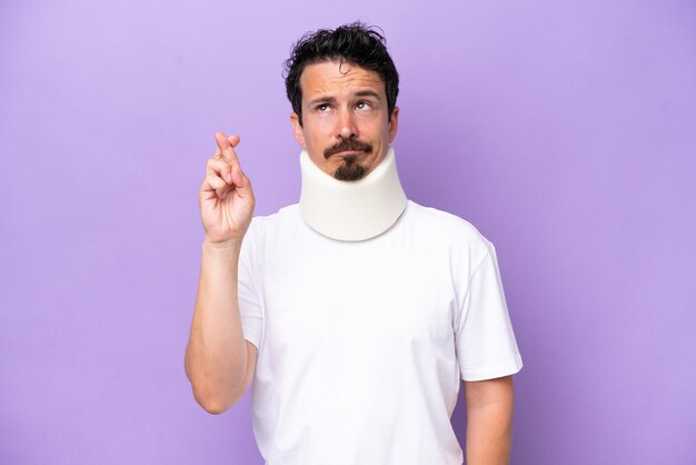 Young caucasian man wearing neck brace isolated on purple background with fingers crossing and wishing the best