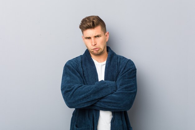 Young caucasian man wearing pajama frowning face in displeasure, keeps arms folded.