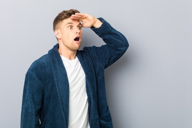 Young caucasian man wearing pajama looking far away keeping hand on forehead.