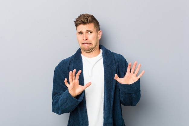 Young caucasian man wearing pajama rejecting someone showing a gesture of disgust.