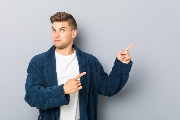 Young caucasian man wearing pajama shocked pointing with index fingers to a copy space.