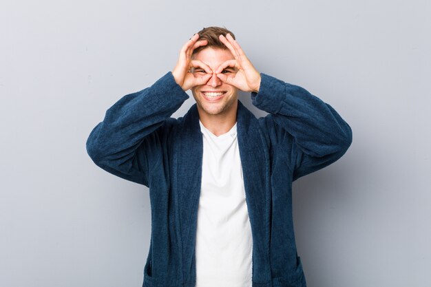 Young caucasian man wearing pajama showing okay sign over eyes