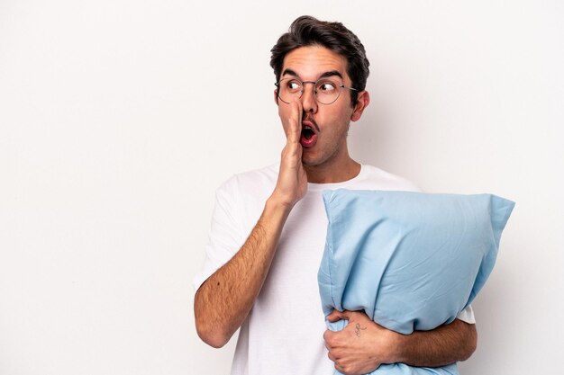 Young caucasian man wearing pajamas holding a pillow isolated on white background is saying a secret hot braking news and looking aside