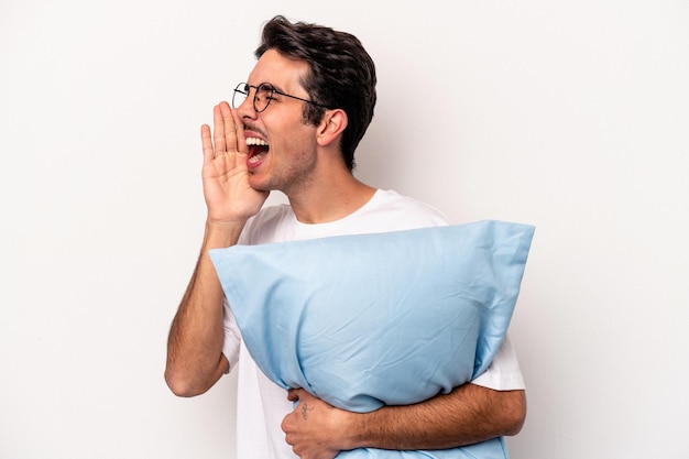 Young caucasian man wearing pajamas holding a pillow isolated on white background shouting and holding palm near opened mouth