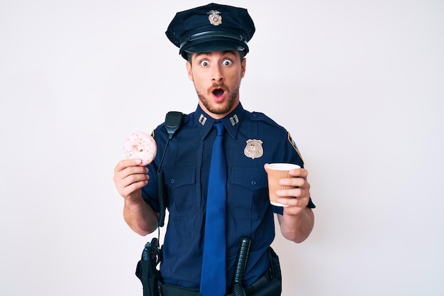 Young caucasian man wearing police uniform holding take away coffee and donut afraid and shocked with surprise and amazed expression, fear and excited face.