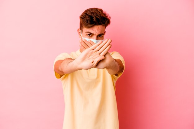 Young caucasian man wearing a protection for coronavirus isolated on pink background doing a denial gesture