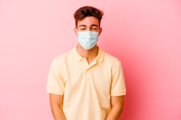 Young caucasian man wearing a protection for coronavirus isolated on pink background laughs and closes eyes, feels relaxed and happy.