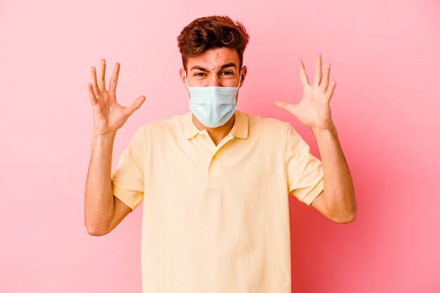 Young caucasian man wearing a protection for coronavirus isolated on pink background screaming with rage.