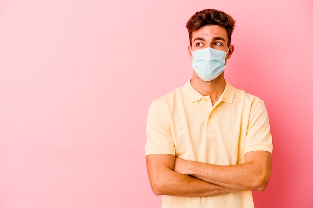 Young caucasian man wearing a protection for coronavirus isolated on pink background smiling confident with crossed arms.