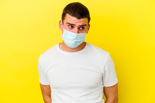 Young caucasian man wearing a protection for coronavirus isolated on yellow background confused, feels doubtful and unsure.