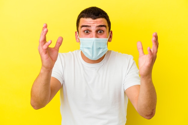 Young caucasian man wearing a protection for coronavirus isolated on yellow background receiving a pleasant surprise, excited and raising hands.