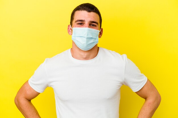 Young caucasian man wearing a protection for coronavirus isolated on yellow wall confident keeping hands on hips.