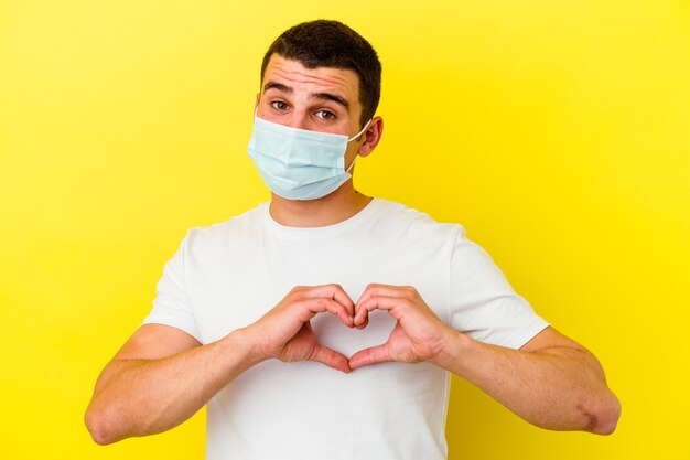 Young caucasian man wearing a protection for coronavirus isolated on yellow wall smiling and showing a heart shape with hands.