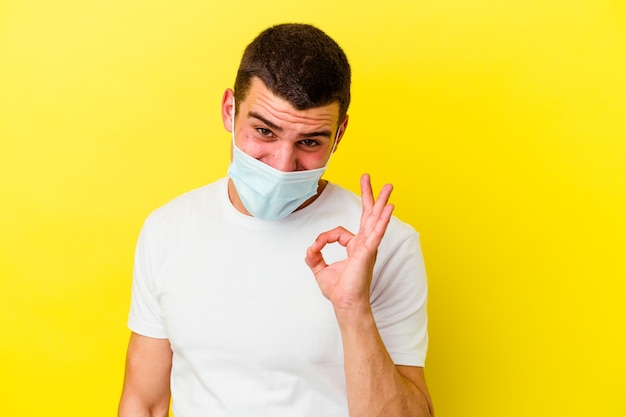 Young caucasian man wearing a protection for coronavirus isolated on yellow wall winks an eye and holds an okay gesture with hand