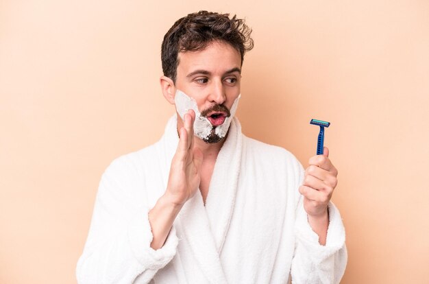 Young caucasian man wearing shaving foam and holding razor blade isolated on beige background is saying a secret hot braking news and looking aside