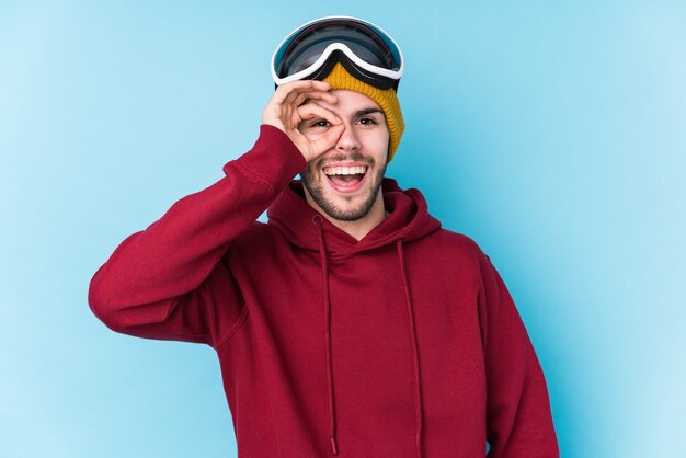 Young caucasian man wearing a ski clothes isolated excited keeping ok gesture on eye.