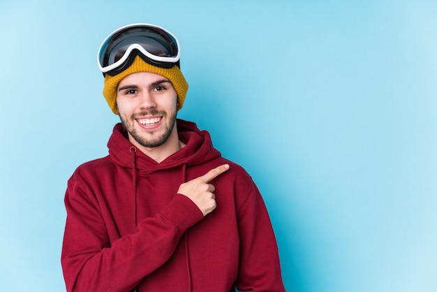 Young caucasian man wearing a ski clothes isolated smiling and pointing aside, showing something at blank space.