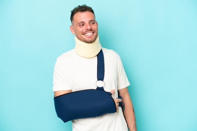 Young caucasian man wearing a sling and neck lace isolated on blue background looking side