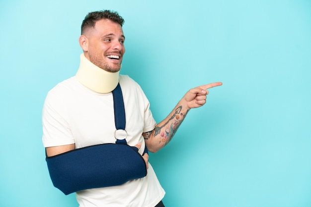 Young caucasian man wearing a sling and neck lace isolated on blue background pointing finger to the side and presenting a product