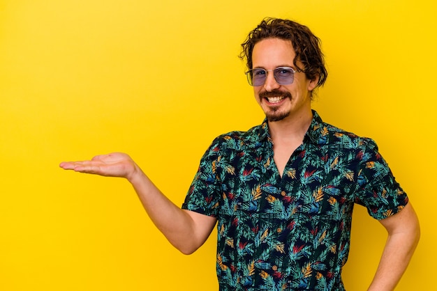 Young caucasian man wearing summer clothes isolated on yellow background showing a copy space on a palm and holding another hand on waist.