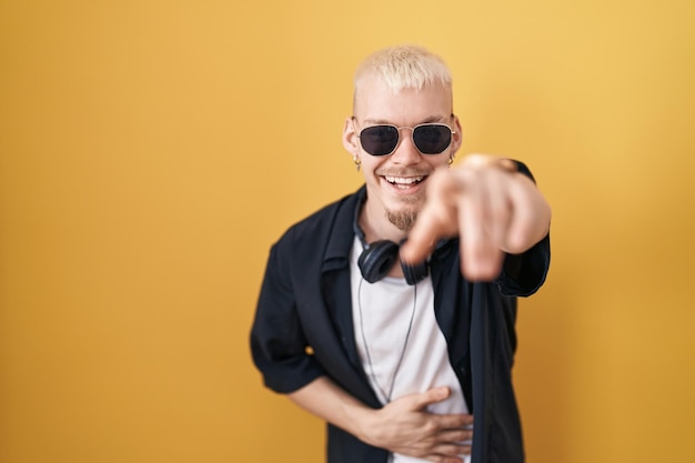 Young caucasian man wearing sunglasses standing over yellow background laughing at you, pointing finger to the camera with hand over body, shame expression