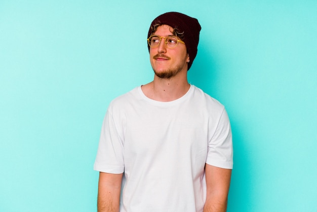 Young caucasian man wearing a wool hat isolated on blue wall  dreaming of achieving goals and purposes