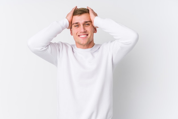 Young caucasian man on white wall laughs joyfully keeping hands on head. Happiness concept.