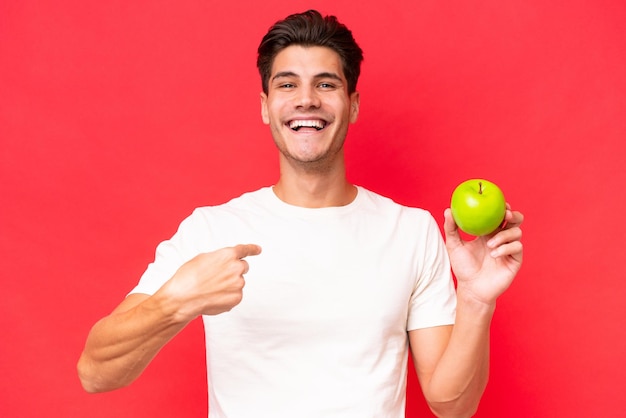 Young caucasian man with an apple isolated on red background with surprise facial expression