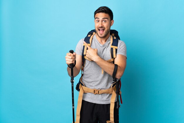 Young caucasian man with backpack and trekking poles isolated on blue background surprised and pointing side