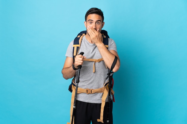Young caucasian man with backpack and trekking poles isolated on blue covering mouth with hands