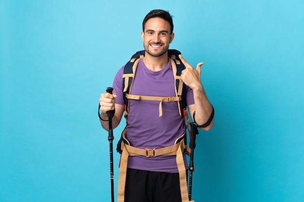 Young caucasian man with backpack and trekking poles isolated on blue  giving a thumbs up gesture