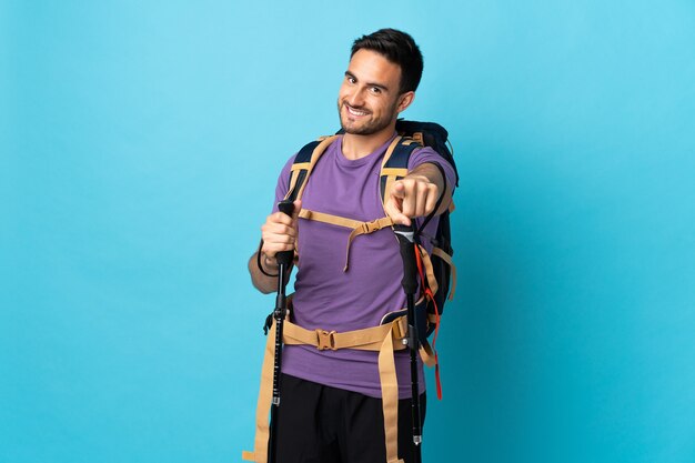 Young caucasian man with backpack and trekking poles isolated on blue wall pointing front with happy expression