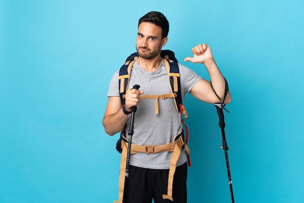 Young caucasian man with backpack and trekking poles isolated on blue wall proud and self-satisfied