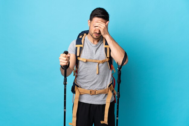 Young caucasian man with backpack and trekking poles isolated on blue wall with headache