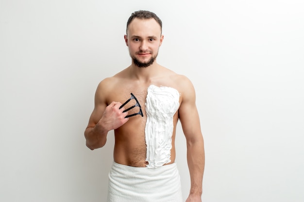 Photo young caucasian man with beard holds razor shaves his chest with white shaving foam man shaving his torso