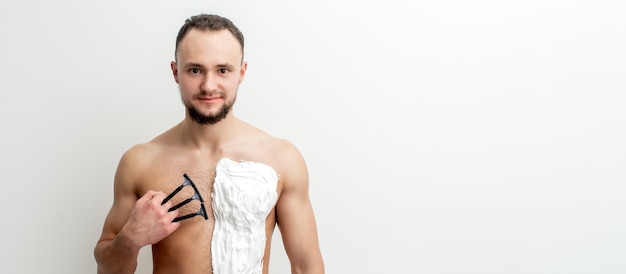 Young caucasian man with beard holds razor shaves his chest with white shaving foam. Man shaving his torso