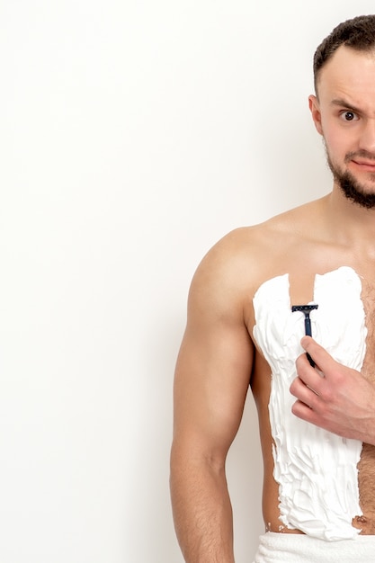 Photo young caucasian man with beard holds razor shaves his chest with white shaving foam on white wall