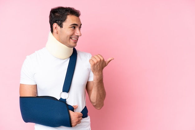 Young caucasian man with broken arm and wearing a sling isolated on pink background pointing to the side to present a product