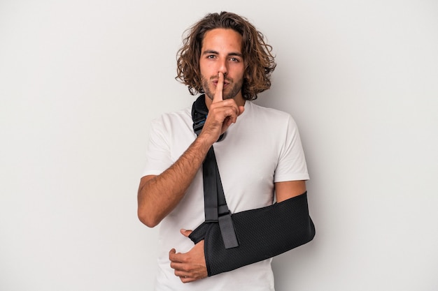 Young caucasian man with broken hand isolated on gray\
background keeping a secret or asking for silence.