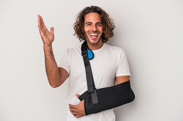 Young caucasian man with broken hand isolated on gray background receiving a pleasant surprise, excited and raising hands.