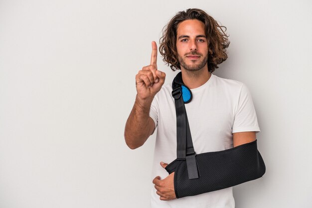 Young caucasian man with broken hand isolated on gray background showing number one with finger.