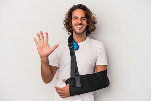 Young caucasian man with broken hand isolated on gray background smiling cheerful showing number five with fingers.