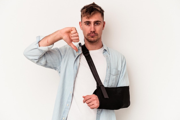 Young caucasian man with broken hand isolated on white background showing a dislike gesture, thumbs down. Disagreement concept.